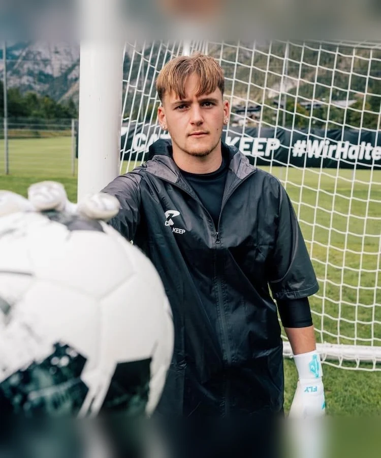 Torwart in schwarzer Jacke mit weißen Fly Soul Torwarthandschuhen und Ball vor dem Tor